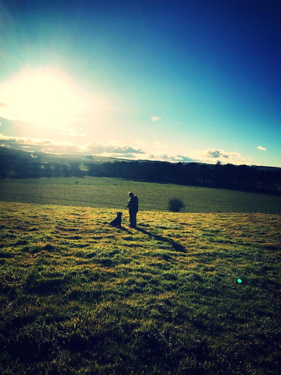 Photo of artists in the distance with her dog, on a hillside with blue sky behind