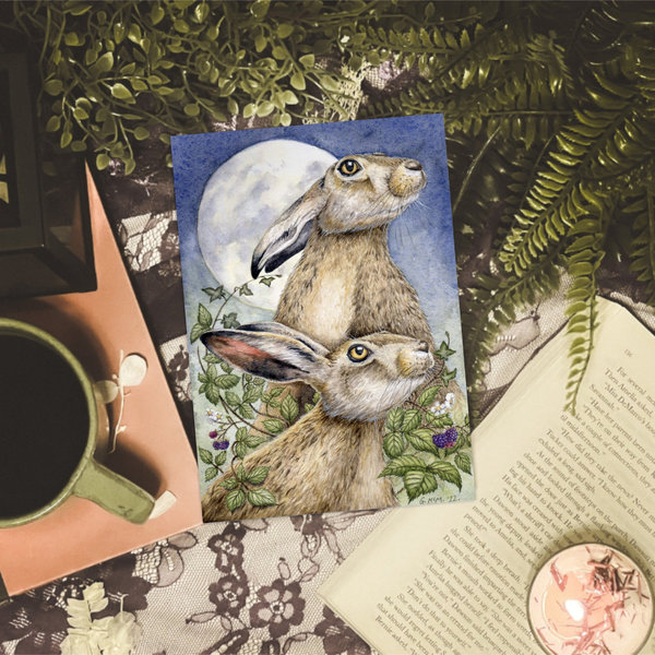 Print of a watercolour painting of hares on a tabletop with mug, plants and book