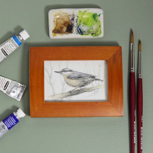 Miniature framed painting of a nuthatch sitting on a green background, surrounded by paints and brushes